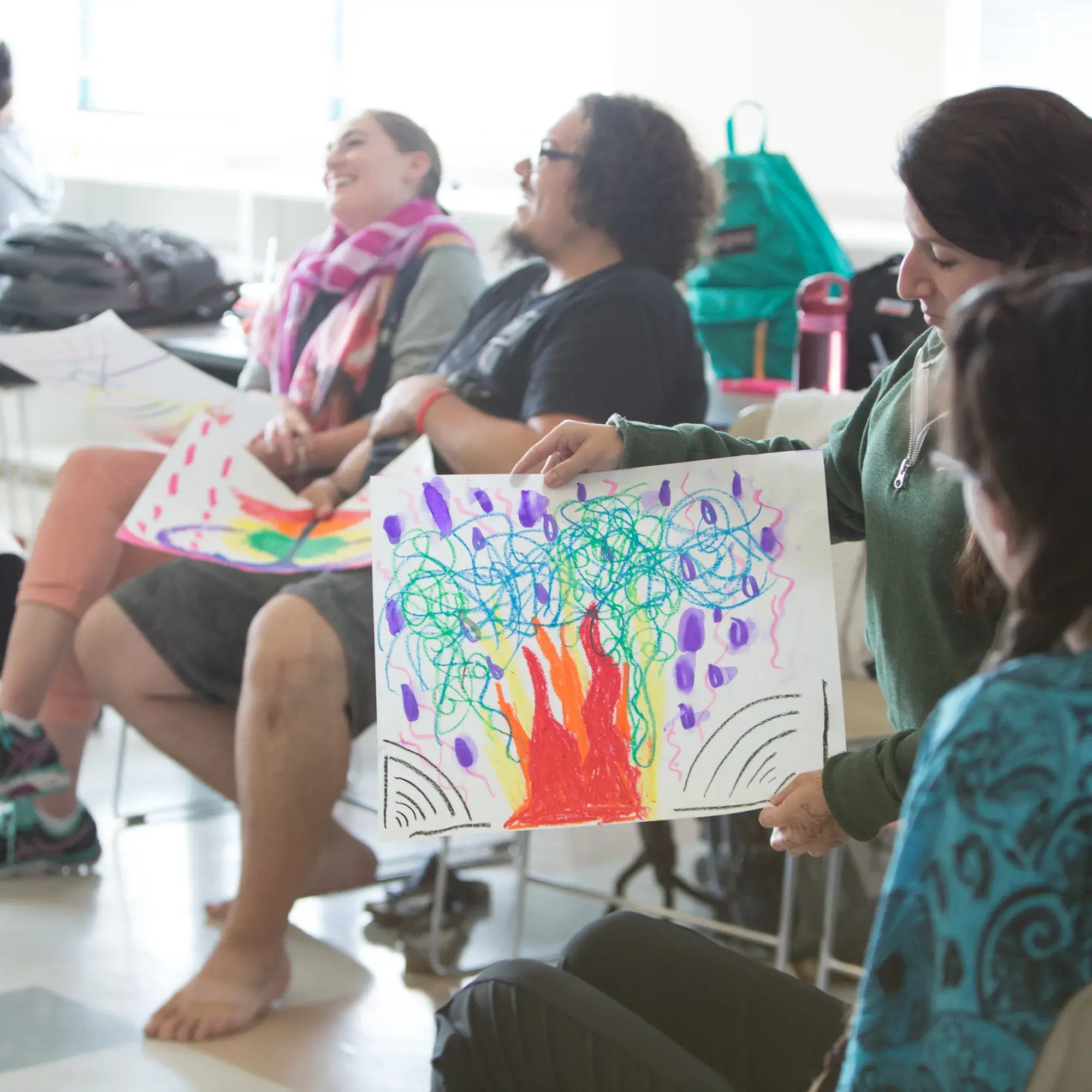 A woman presents her expressive drawing during a group art therapy session. The positive and relaxed reactions of the other participants, who are smiling and holding their own artwork, highlight a moment of creative sharing and connection.