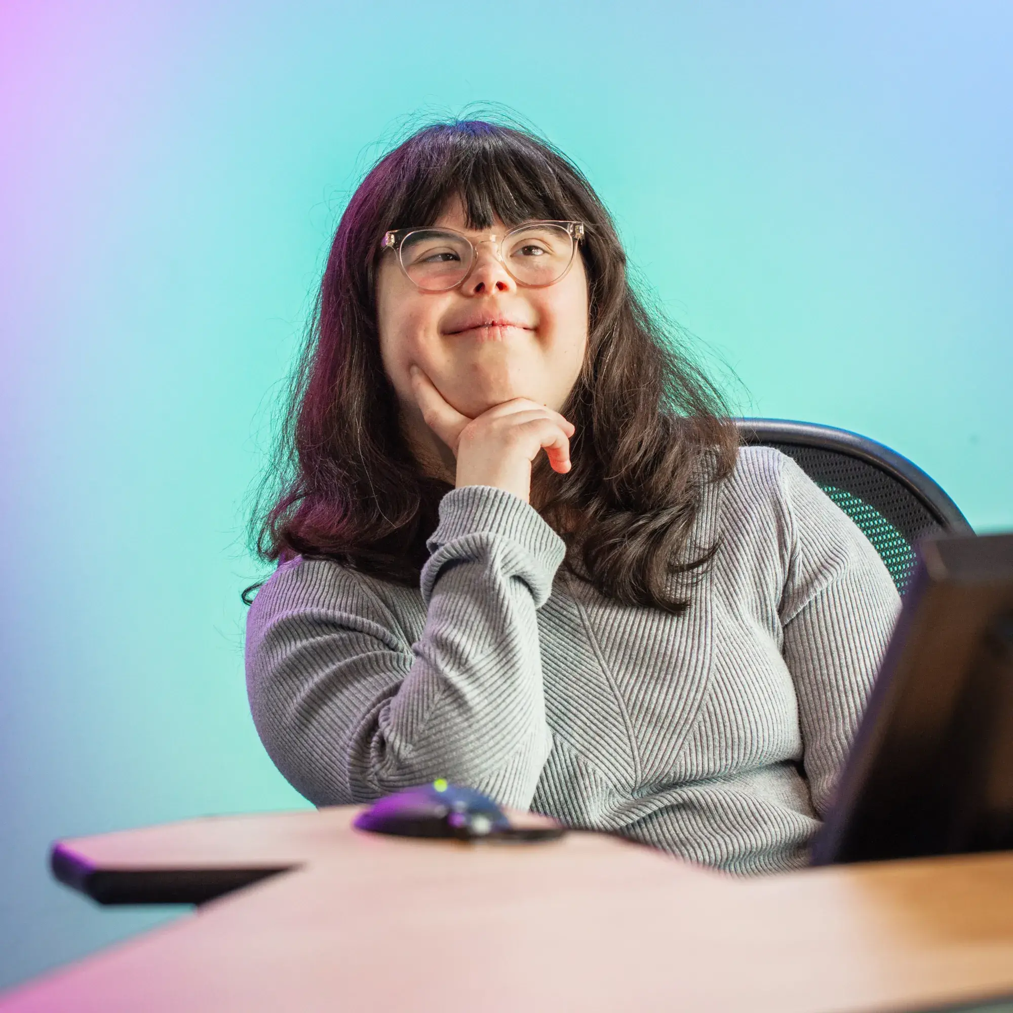 A young woman with long dark hair and glasses sits confidently at a uniquely shaped wooden desk, her chin resting on her hand in a thoughtful pose. She wears a ribbed, light gray long-sleeve shirt and smiles subtly while gazing slightly to the side. Behind her, a softly lit background fades from pink and purple on the left to turquoise and blue on the right, creating a vibrant, creative atmosphere. A computer monitor and mouse are visible in the foreground.