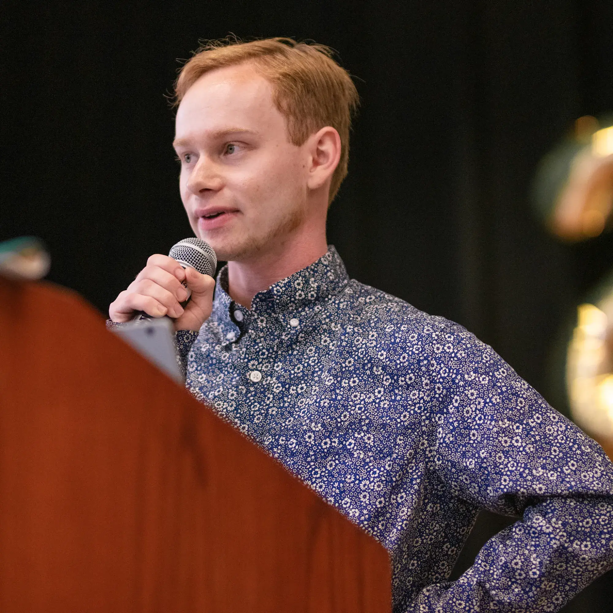 A young man with light brown hair stands at a podium, holding a microphone and speaking. He wears a dark blue button-up shirt with a white floral pattern. A teal cloth drapes over the edge of the podium, and the background is dark with blurred gold balloons partially visible.