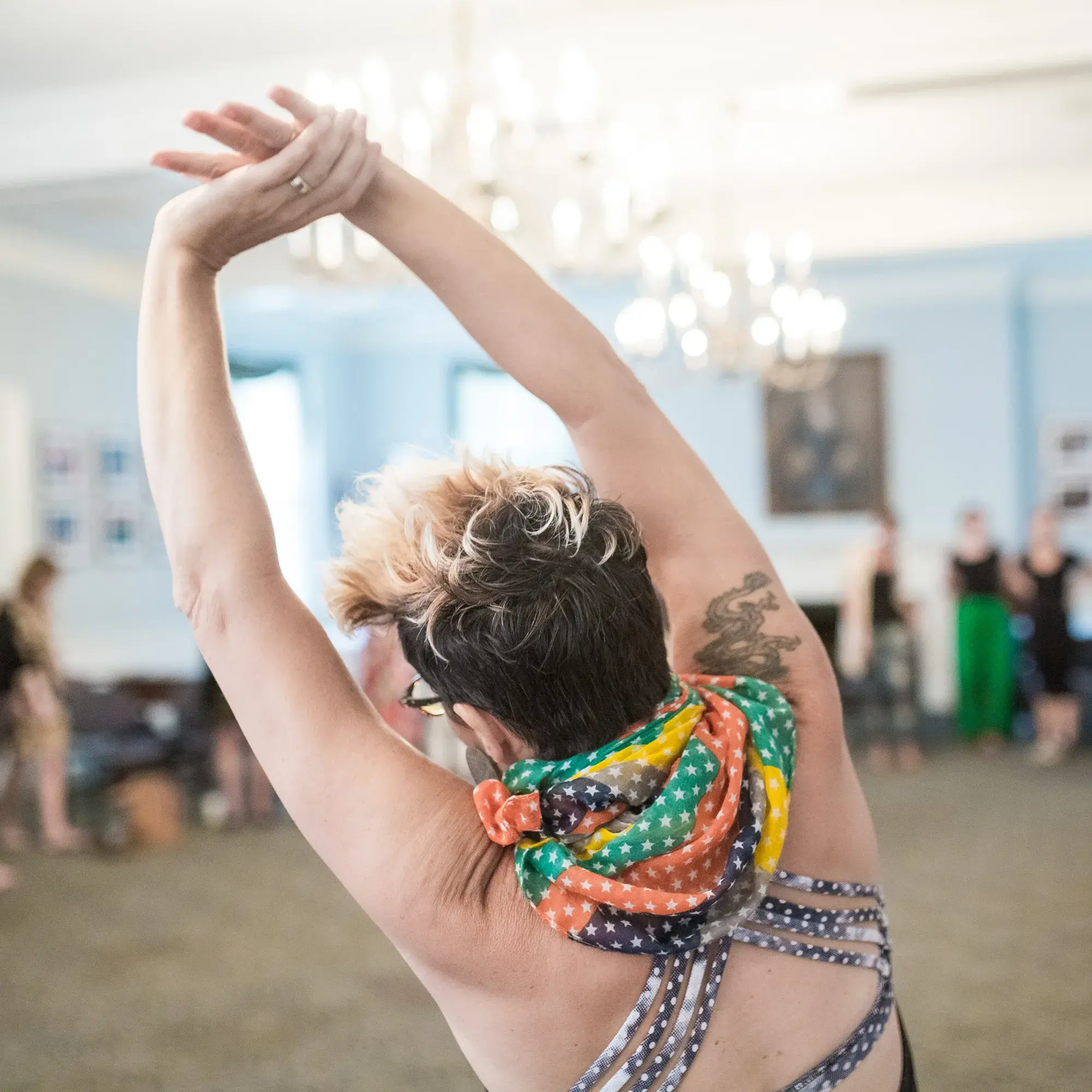 A participant in a mindful movement workshop performs a deep side stretch. The close-up shot from behind focuses on their raised arms and colorful scarf, with the rest of the class and the chandeliers of the room softly blurred in the background.