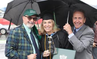 Anusia Hirsch wears her graduation robe and is stading with advisor Gene Ferraro and her husabnd under umbrellas holding her award and diploma