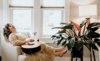 Lauren Pratt laughing in an arm chair with a guitar in her lap and a plant at her feet