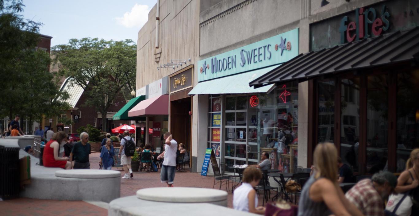 Row of restaurants and shops in Harvard Square. Some people are walking by, some are sitting.