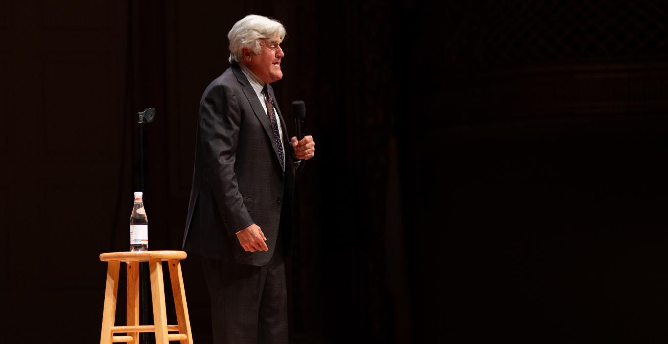 Jay Leno standing on Symphony Hall stage with a stool in the foreground