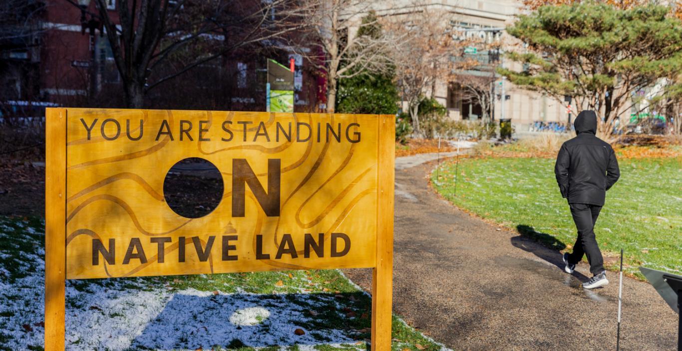 Sign: "You are standing on Native Land" - installed outside, person walking by.