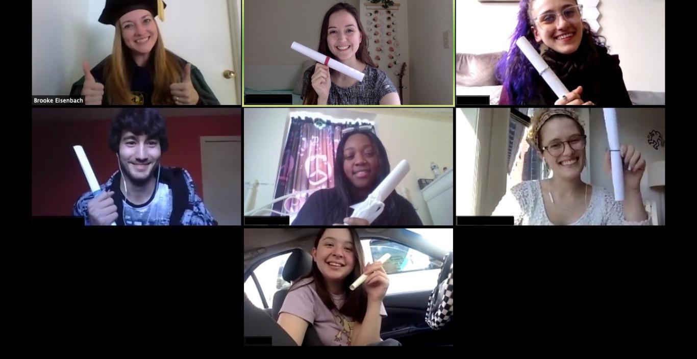 Screenshot of a Zoom call, students hold up paper rolled up to look like diplomas. Professor Eisenbach wears her regalia.