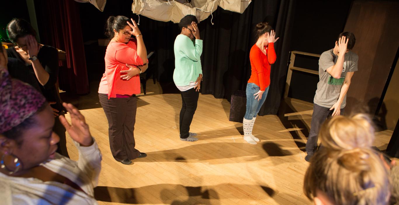People in a circle on a stage during a drama therapy exercise stand with one hand up against their faces.