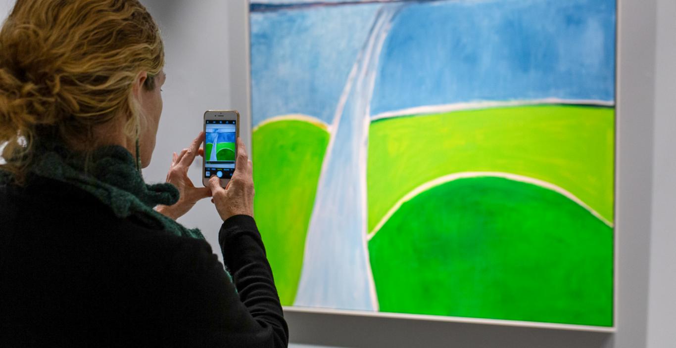 A woman takes a photo of a painting with her cell phone.