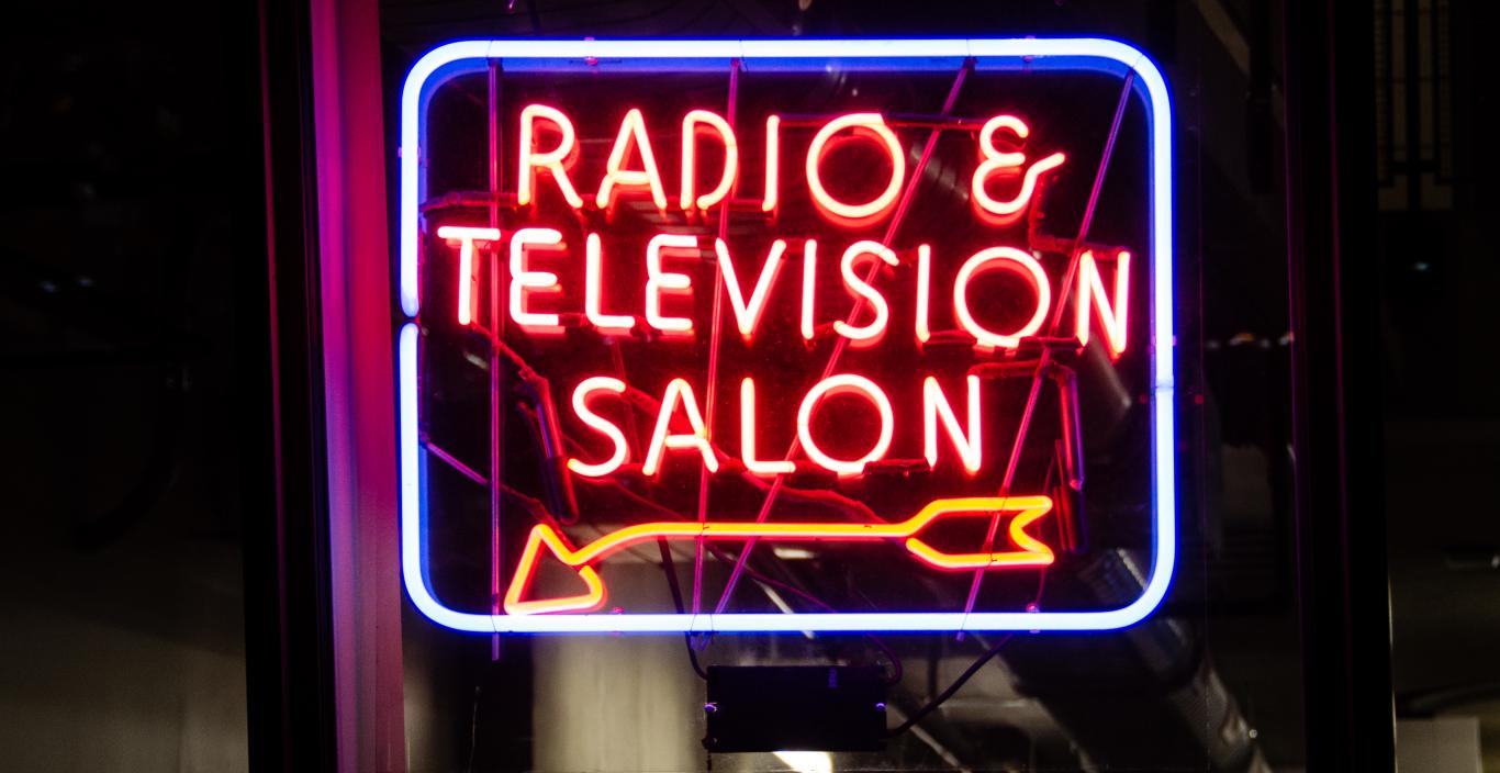 Neon sign that reads: "radio and television station" with an arrow pointing to the bottom left corner of the rectangular sign.