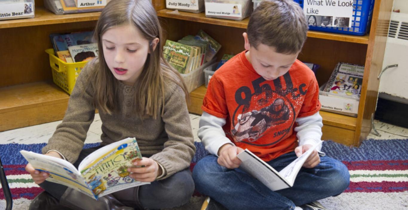 A girl and boy are sitting on the floor reading in the book corner.