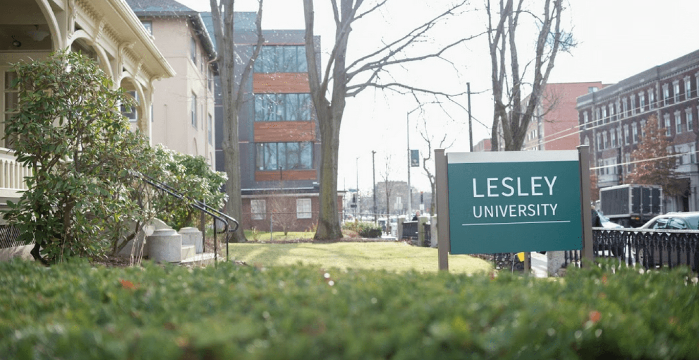 Photograph of admissions building shrubs and exterior with Lesley sign and sidewalk in the background.