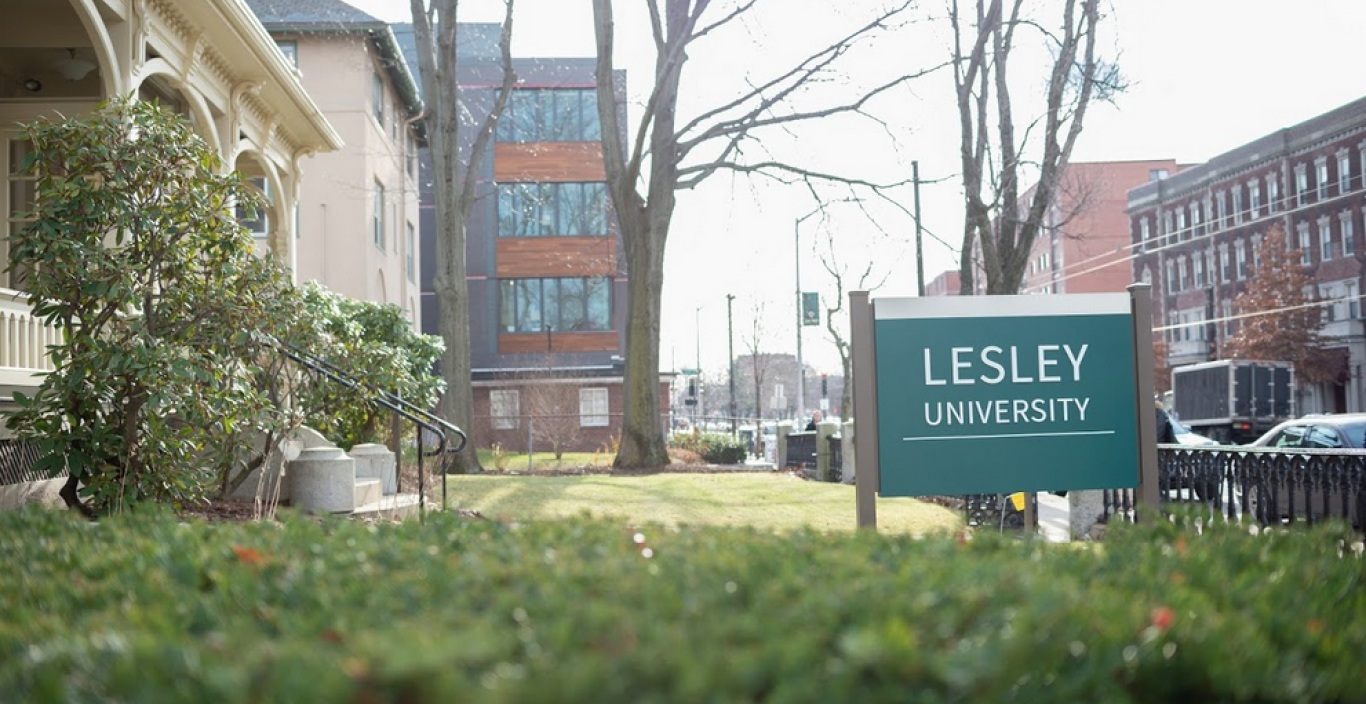 Photograph of admissions building shrubs and exterior with Lesley sign and sidewalk in the background.