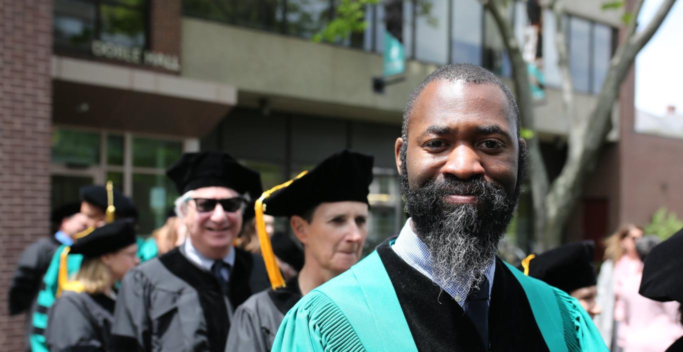 Matthew E. Henry wearing graduation robes with other people in the background. Standing outside.