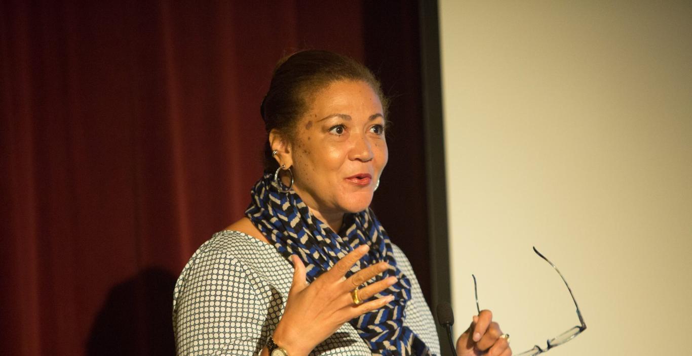 Michele Norris speaks at a podium, holding a pair of glasses in her left hand.