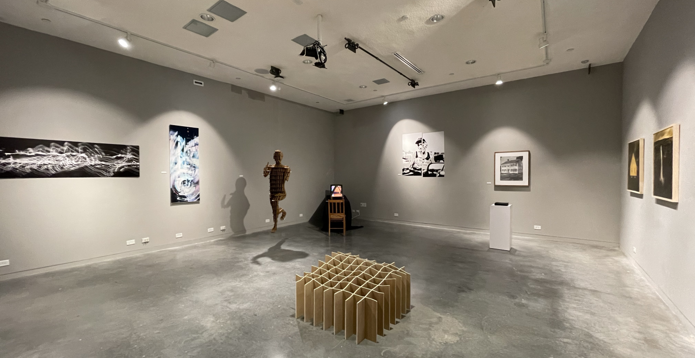 Photo of a gallery exhibit featuring a figurine sculpture and a grid sculpture. The walls and floor are gray. 