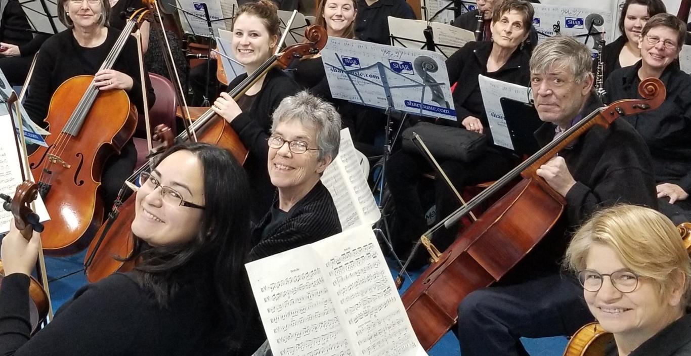 An orchestra seated in with men and women holding instruments including violins, cellos and clarinets.