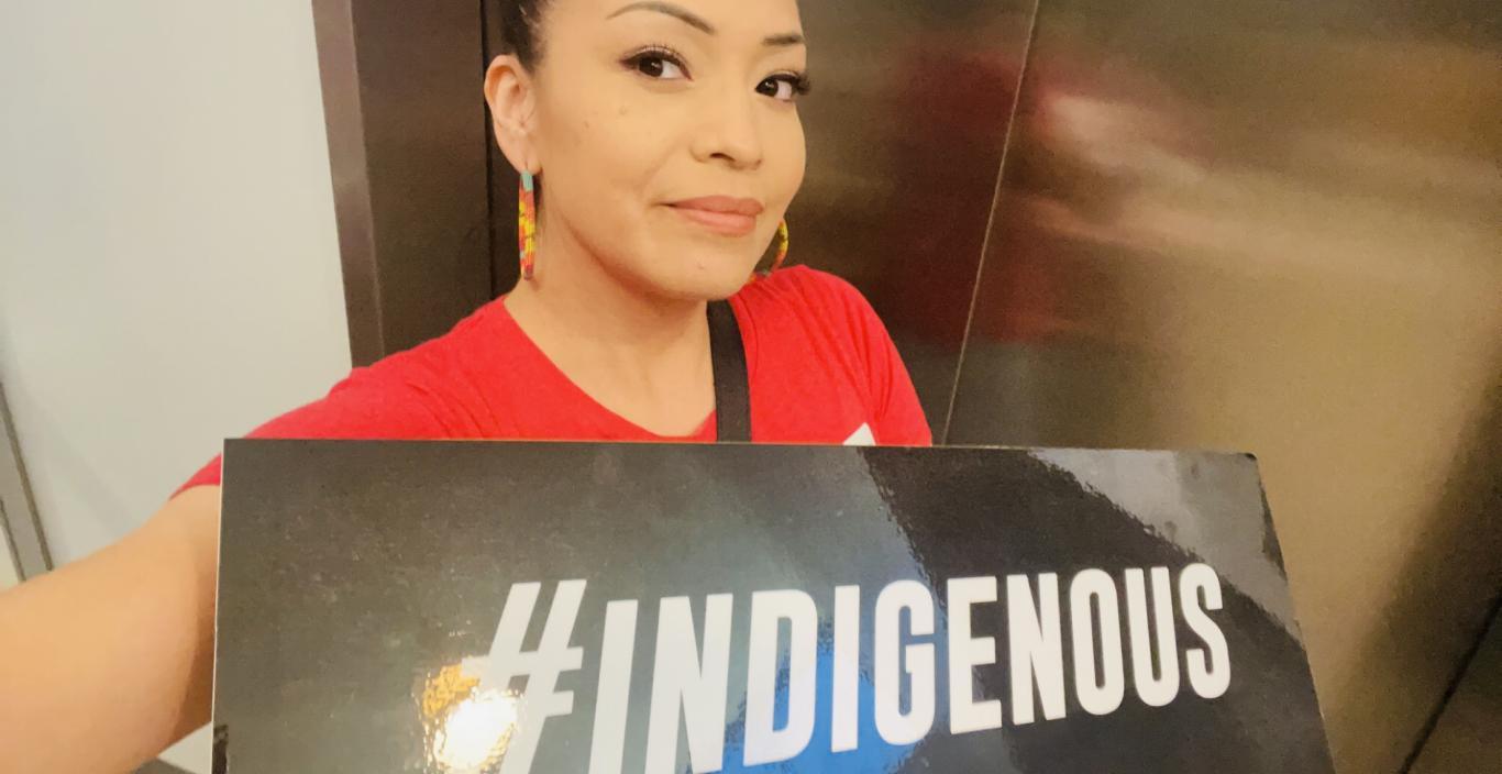 Serene Thin Elk holding a sign that says "#INDIGENOUS"