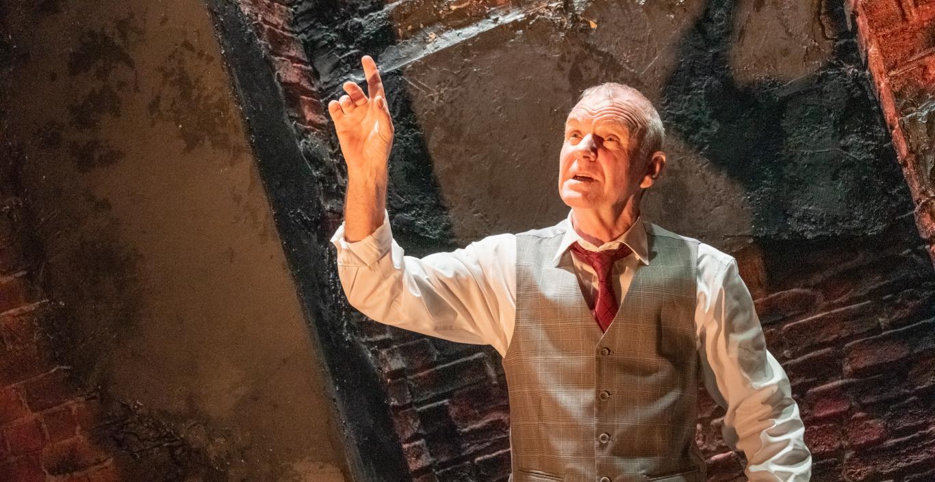 Man in a suit vest and pants holding up one hand, with a brick wall behidn him.