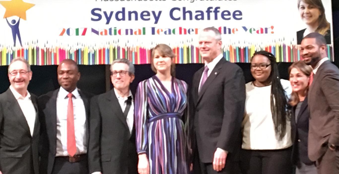 Sydney Chaffee stands on stage with Gov. Charlie Baker, colleagues and students at her ceremony. Behind her is a sign that reads Massachusetts Congratulates Sydney Chaffee.