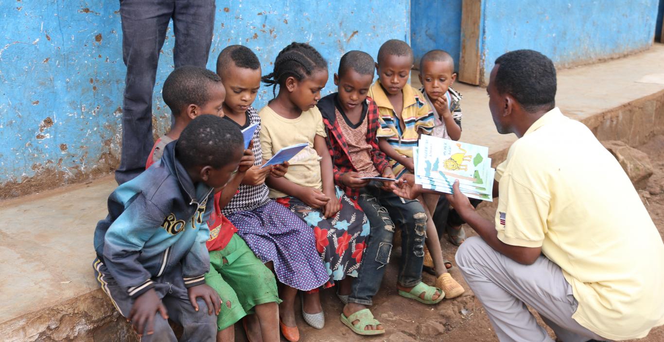 Children in Ethiopia sitting on the sidewalk while their teacher shows them a book.