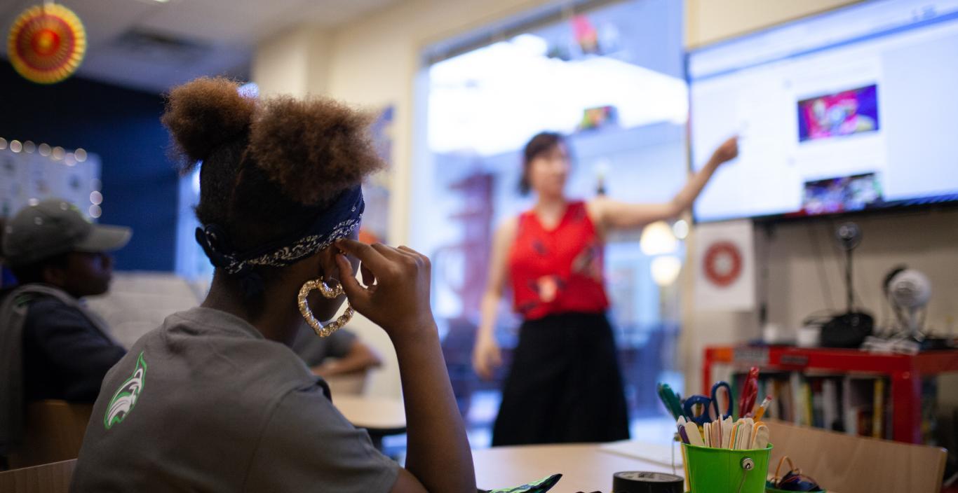 Back of a teenage girl's head with someone teaching in the background.