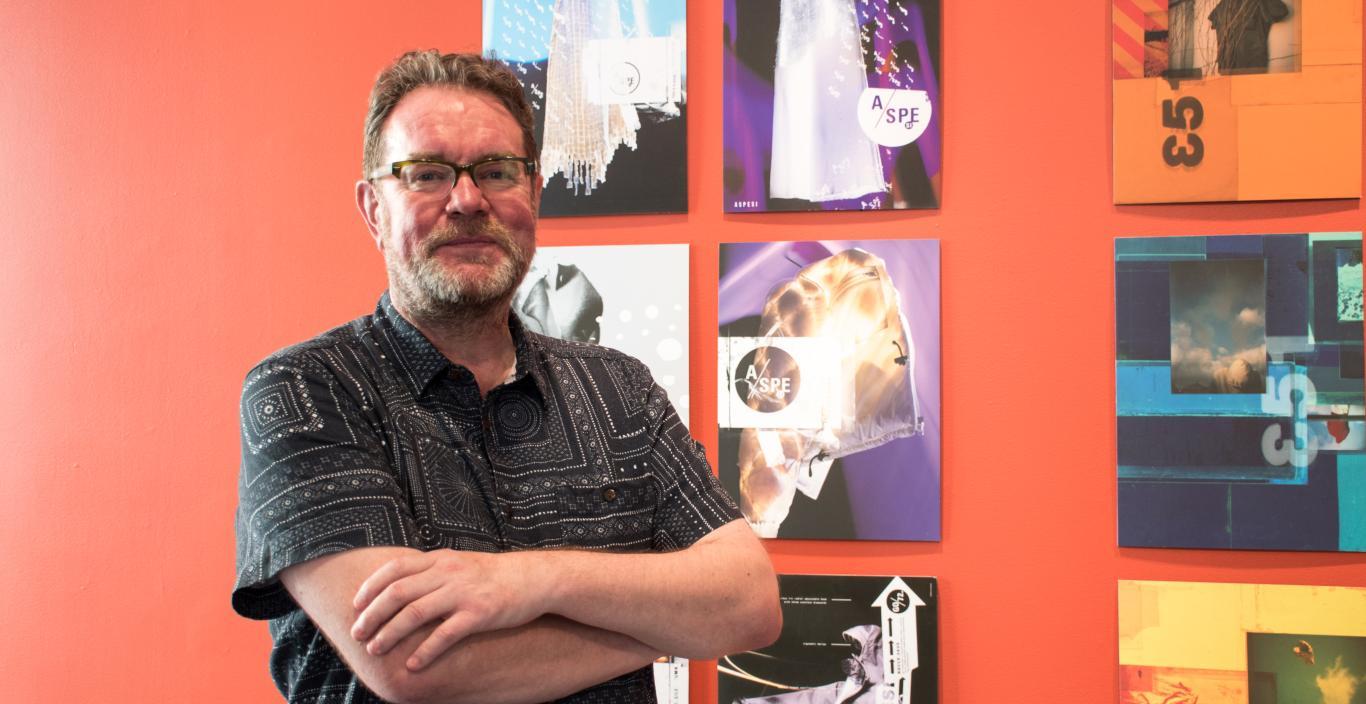 Graphic designer Vaughan Oliver stands with arms crossed and a slight smile in front of posters he's designed.