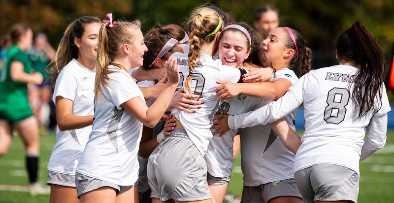 Photograph of members of the Lesley University women's soccer team celebrating on the field in their white jerseys.