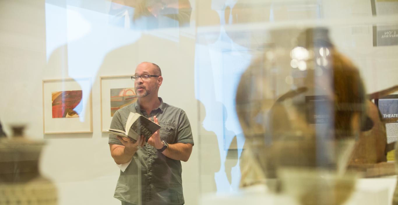 Artsy pick of Aaron Smith reading from a book, taken through a glass case at a museum.