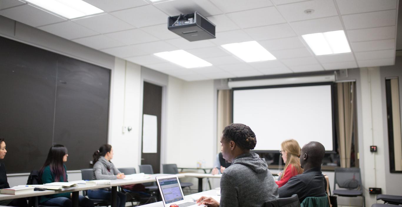Photo of six students sitting in a square shape at tables in a classroom. Main student in focus is engaged in materials on her laptop.