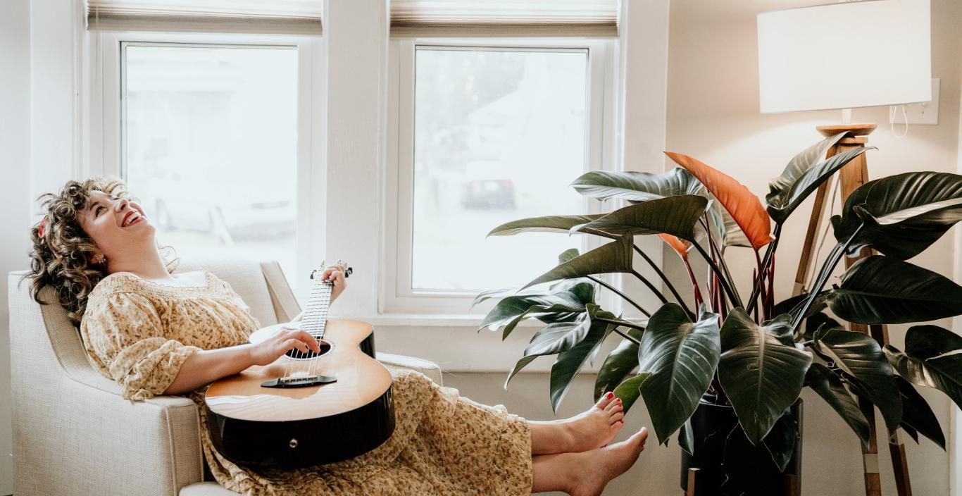 Lauren Pratt laughing in an arm chair with a guitar in her lap and a plant at her feet