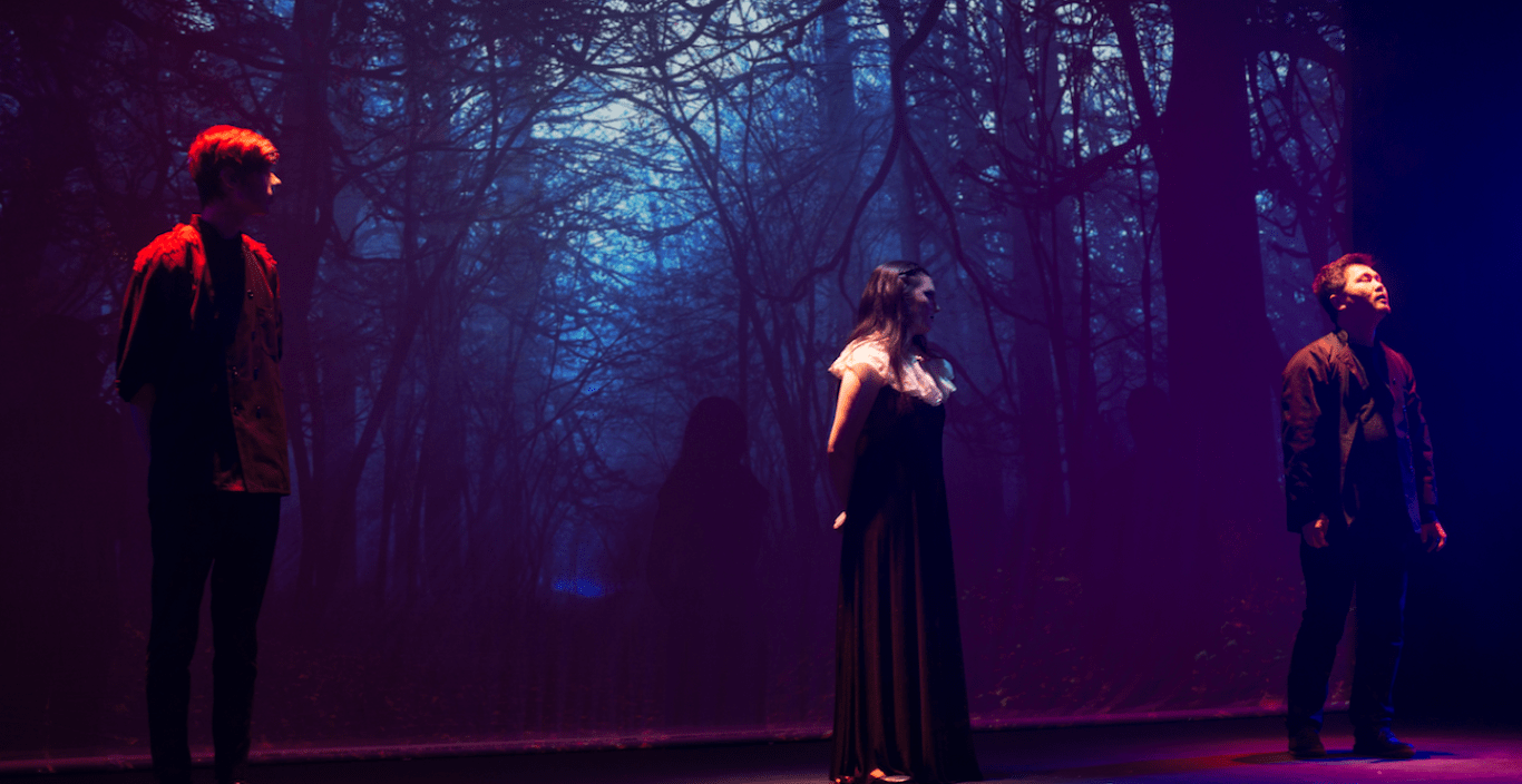 Three actors on stage against the backdrop of the woods in a production of Macbeth