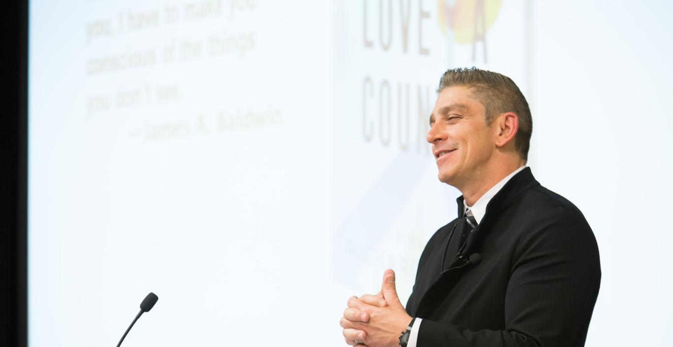 Richard Blanco standing at a podium with hands clasped and smiling.