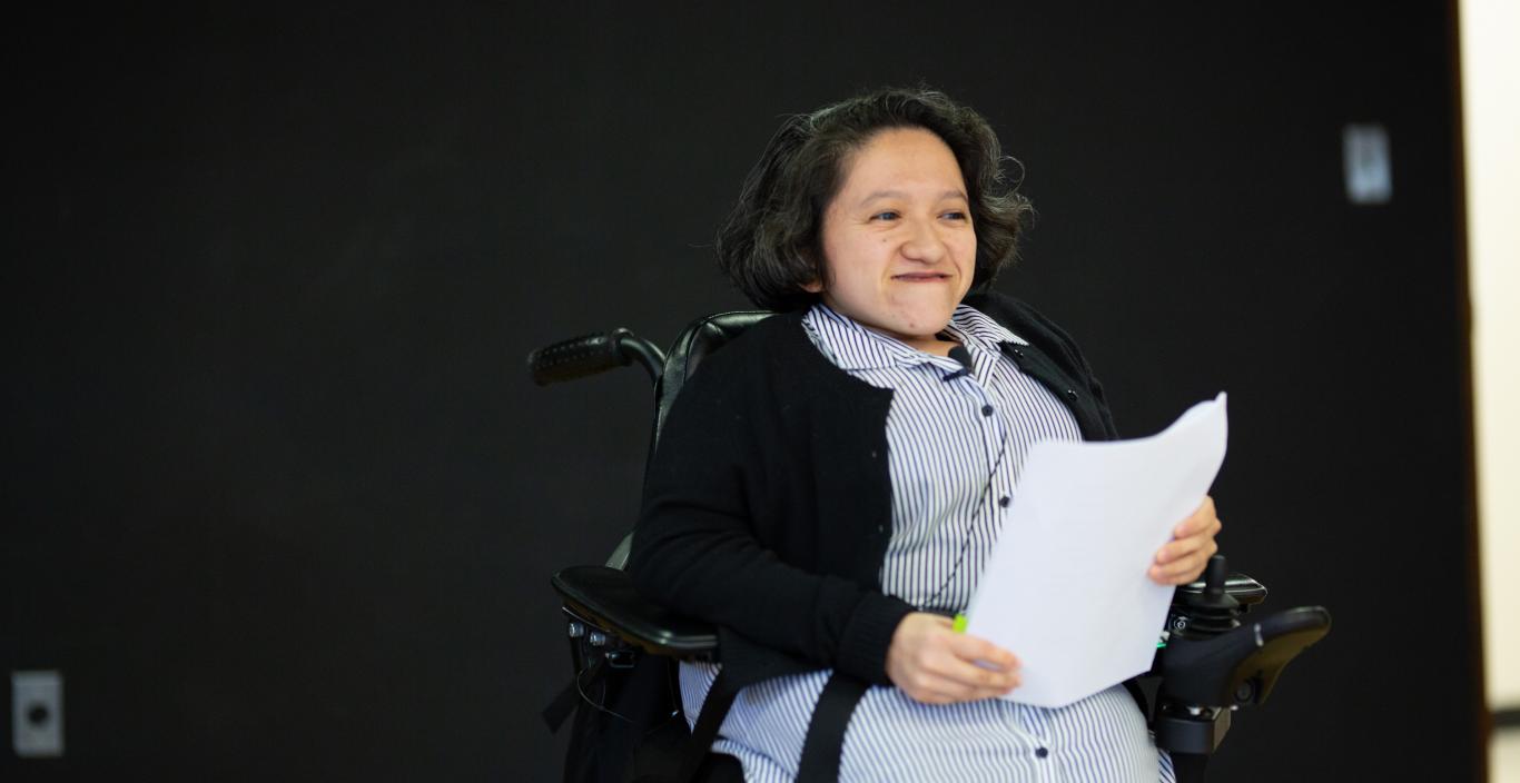 Photograph of young woman in a blue dress and a black sweater seated in a wheelchair holding a piece of paper on a stage.