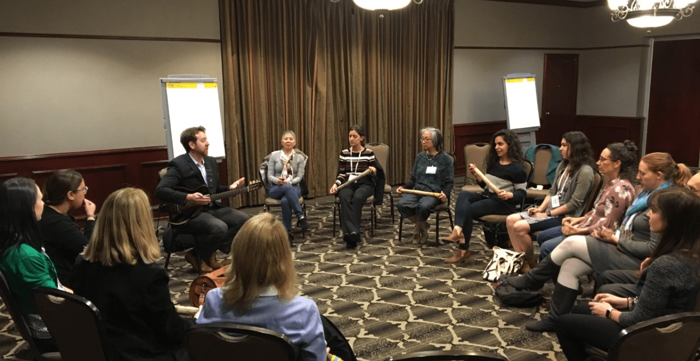 People sitting in a circle in a hotel conference room, Seneca Block holds a guitar and addresses the group