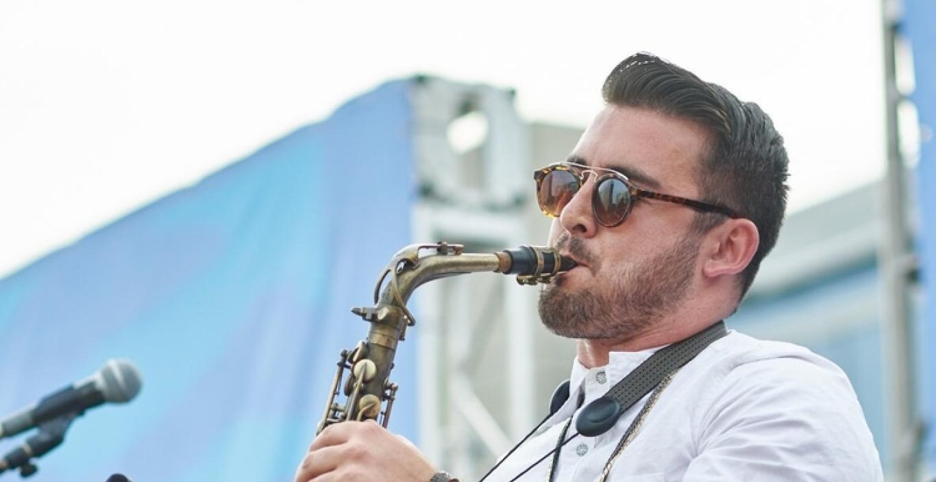 Man with sunglasses playing a saxophone on an outdoor stage.