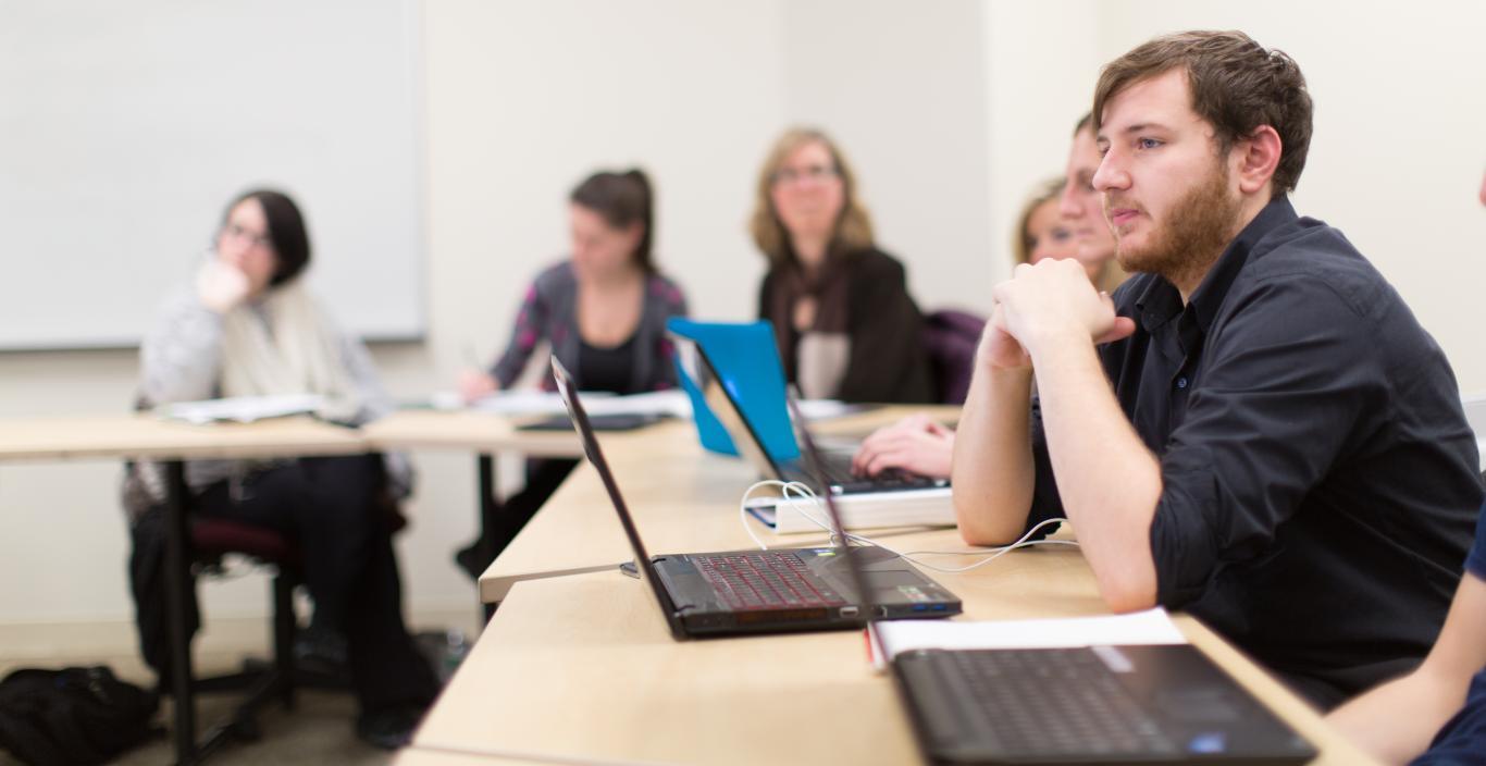 Students in classroom off-campus, with focus on one boy student sitting with hands folded on the table with a laptop in front of him.
