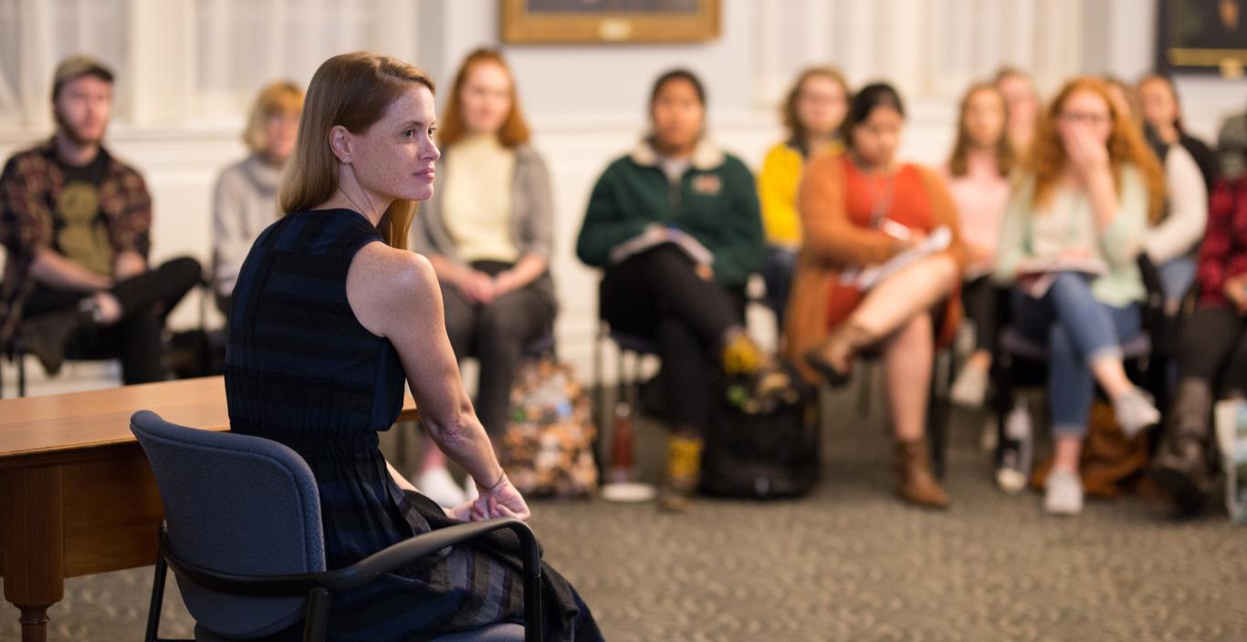 A women with long red hard sitting in a chair speaking to an audience of students