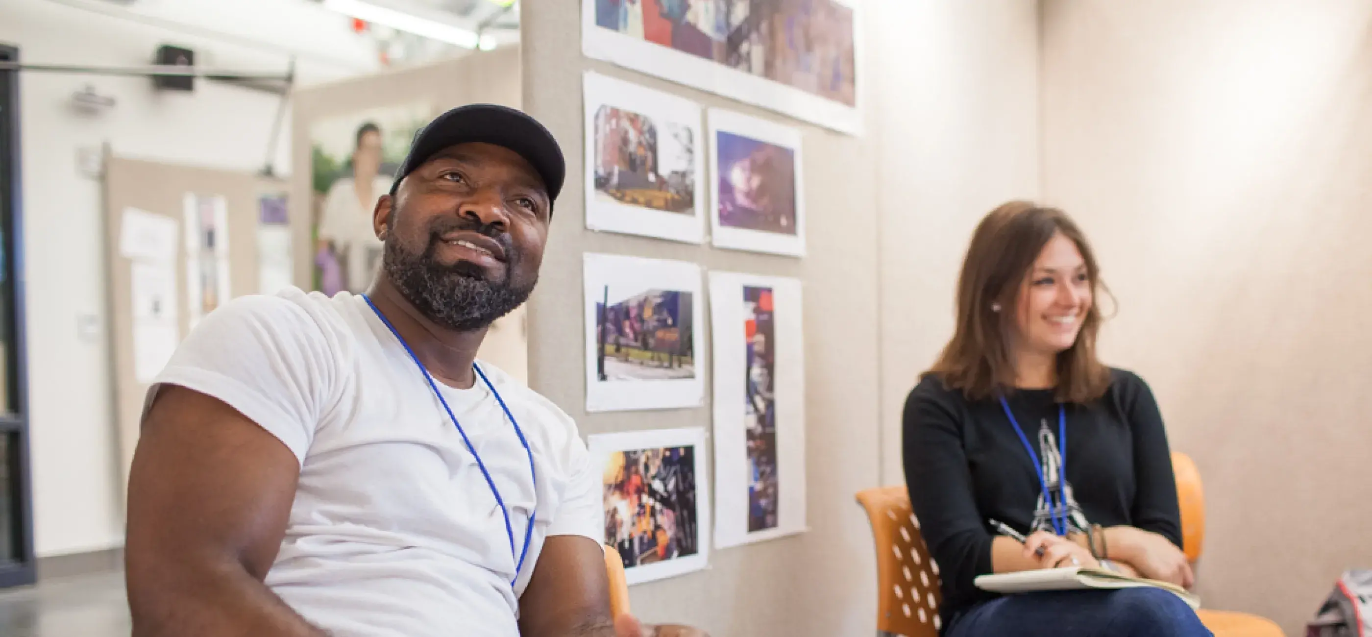 Two adults sit in a classroom or studio setting, smiling and engaged in discussion. The man on the left wears a white t-shirt, jeans, and a black cap, holding a pencil and notebook. The woman on the right is dressed in a black top and jeans, also taking notes. Behind them, artwork or photographs are displayed on a beige wall panel. The setting appears informal and creative, suggesting a workshop or residency environment.