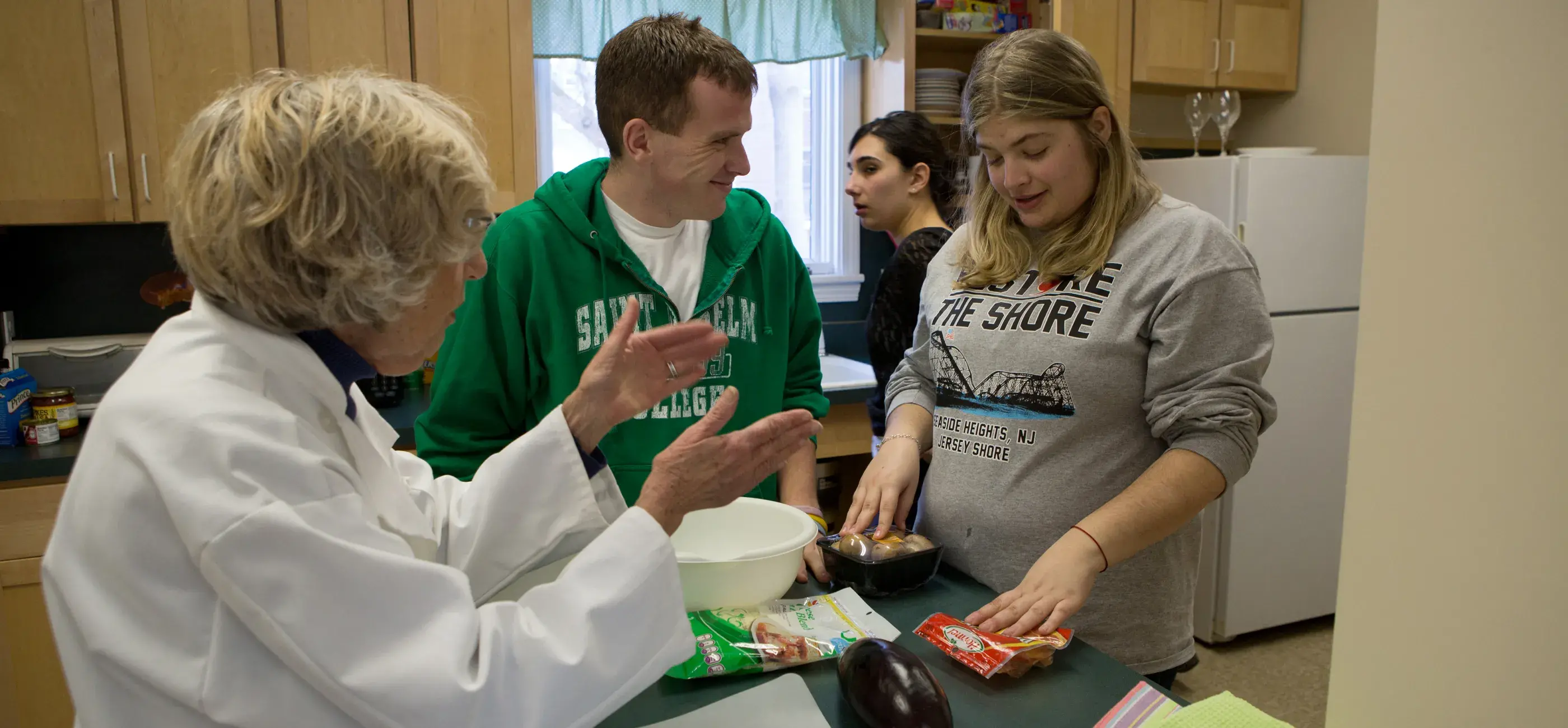 A group of four people are gathered around a kitchen counter in a home-like setting. An older woman in a white coat appears to be instructing or guiding the group, while two young adults listen attentively and one young woman prepares ingredients, including eggplant and packaged food. The background shows wooden cabinets, a refrigerator, and a window with green curtains.