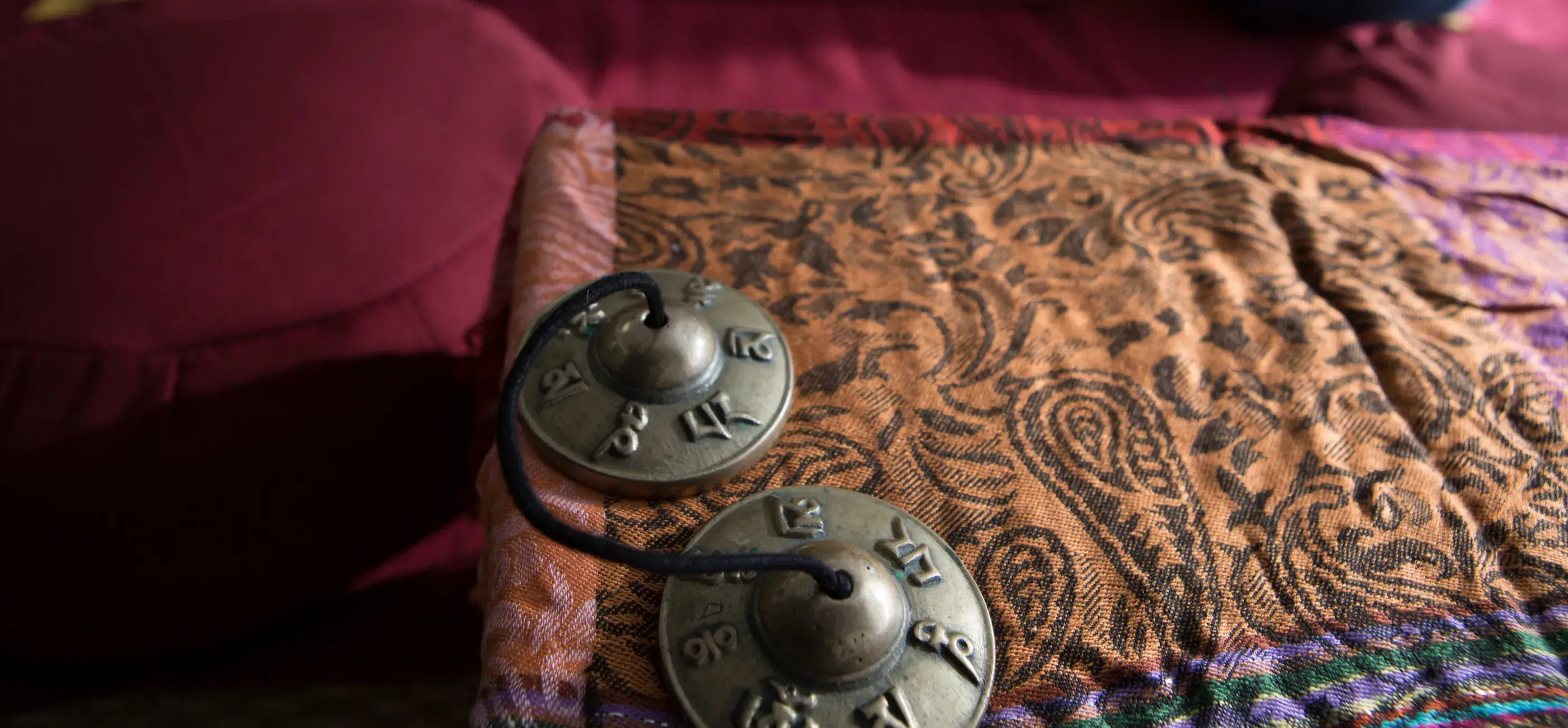 A tranquil meditation space is shown, with a close-up focus on a pair of small, brass cymbals resting on a paisley-patterned fabric. Behind them, soft meditation cushions (zafus) in various colors sit on a red mat, ready for use.