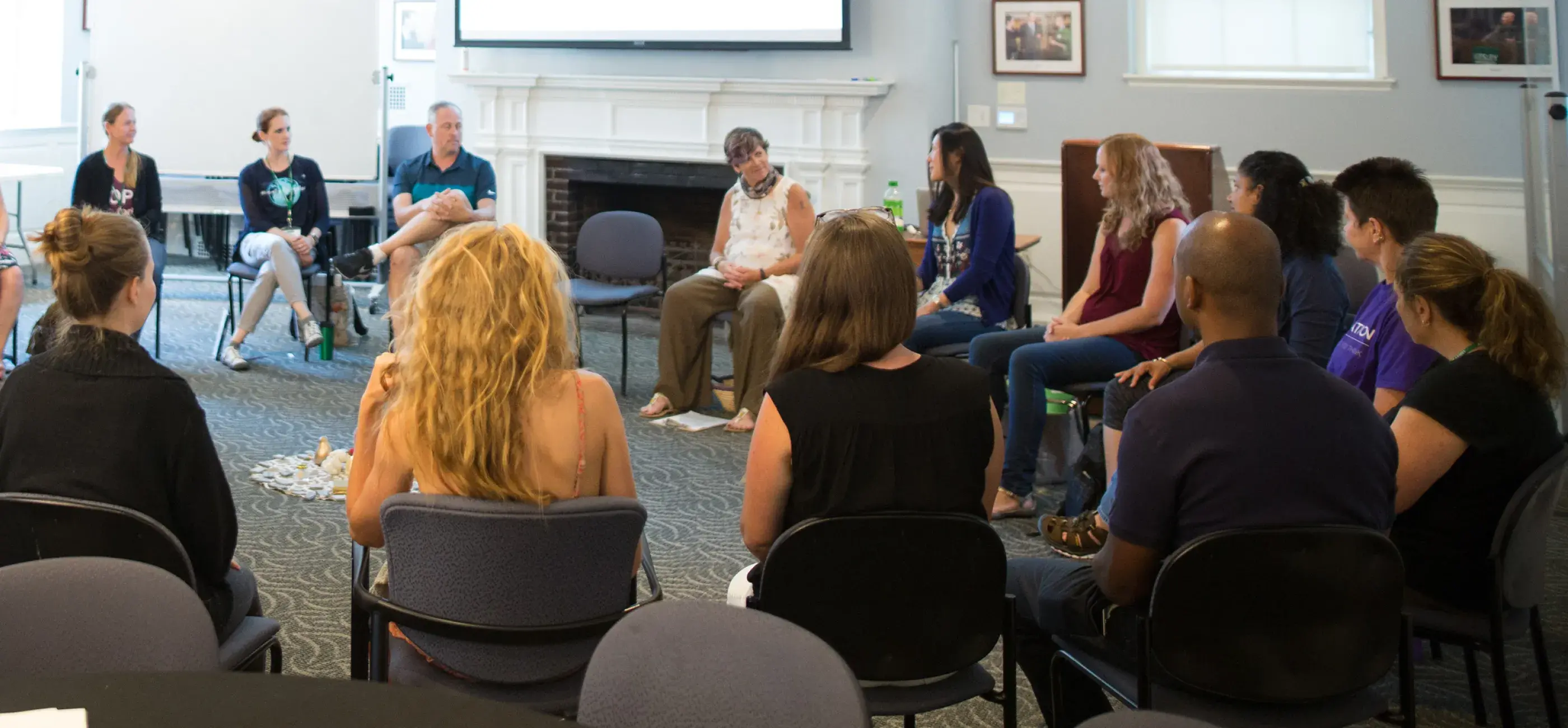 An audience listens attentively during a seminar in a spacious, traditionally decorated hall with a chandelier. The participants are arranged in a large semi-circle, focusing on the presenters and a large screen at the front of the room.