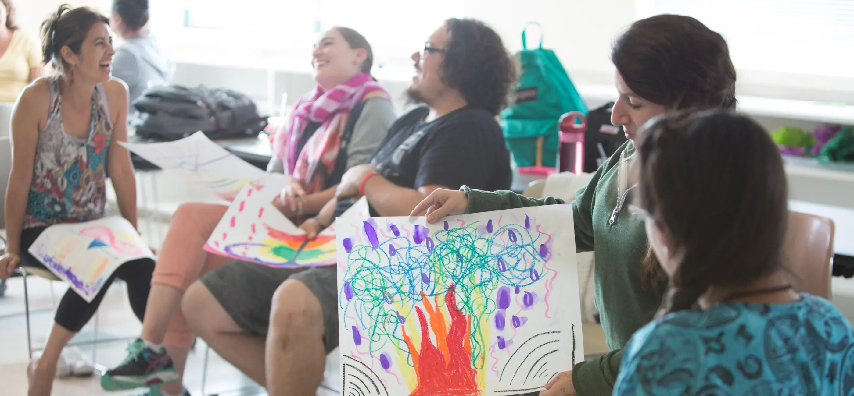 A woman presents her expressive drawing during a group art therapy session. The positive and relaxed reactions of the other participants, who are smiling and holding their own artwork, highlight a moment of creative sharing and connection.