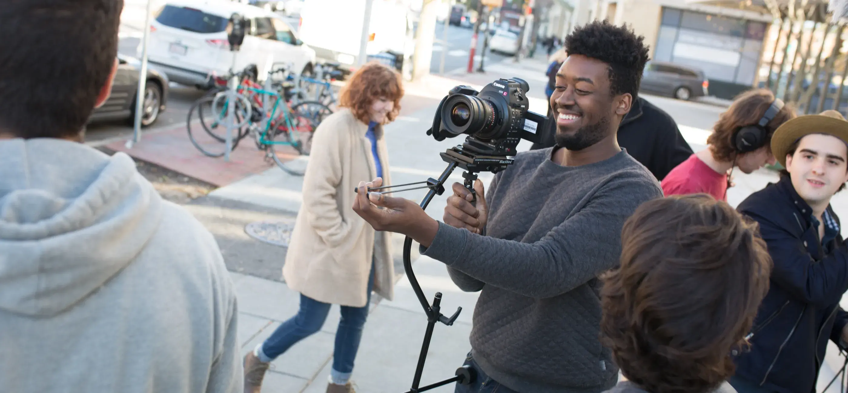 A student film crew captures a scene on a city street. The focus is on the smiling camera operator maneuvering a professional camera rig, highlighting the collaborative and practical nature of filmmaking education.