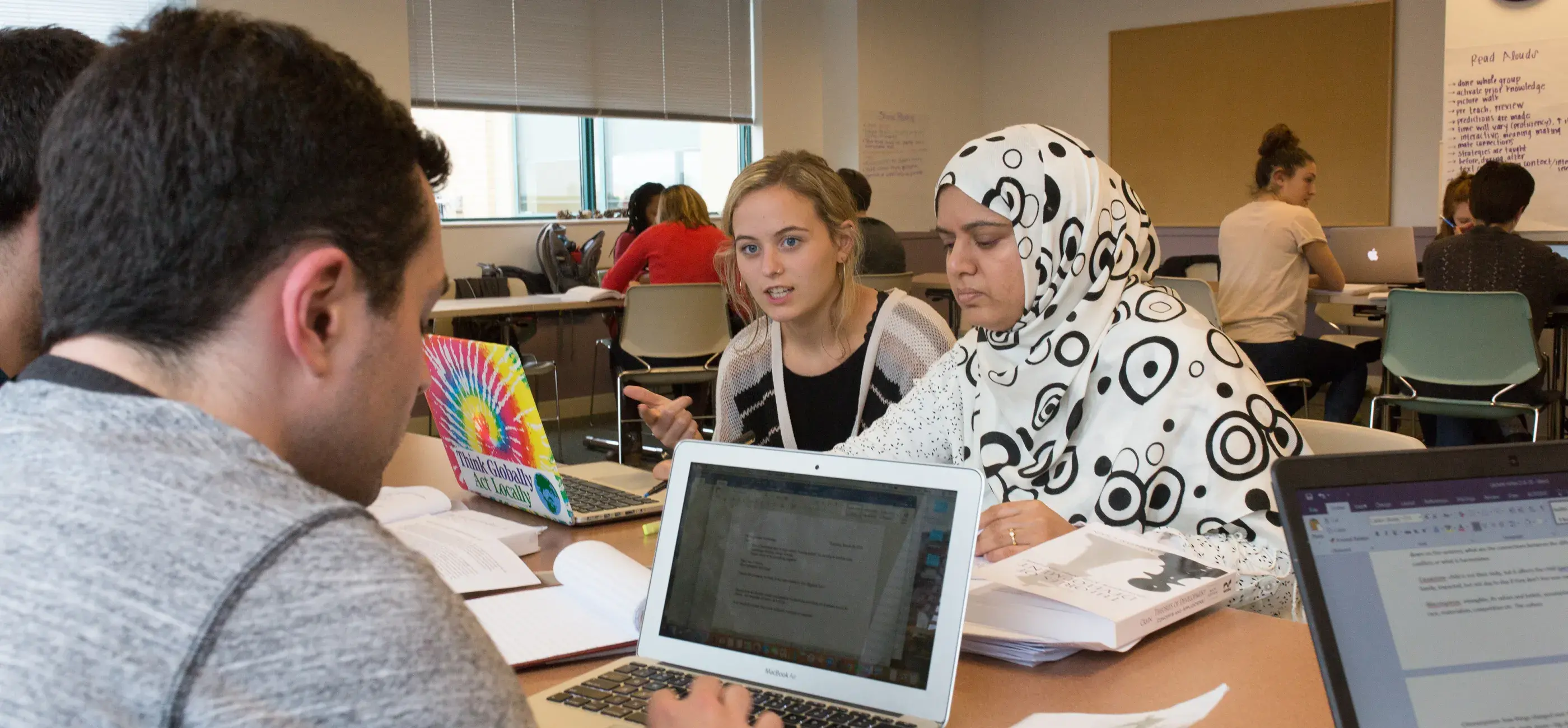 A diverse group of university students collaborates in a classroom, seated around a table with open laptops, notebooks, and textbooks. One student in a patterned hijab is focused on writing, while another student gestures as she speaks. A third student looks at his laptop screen. The classroom is bright, with posters on the wall and other students working in the background.