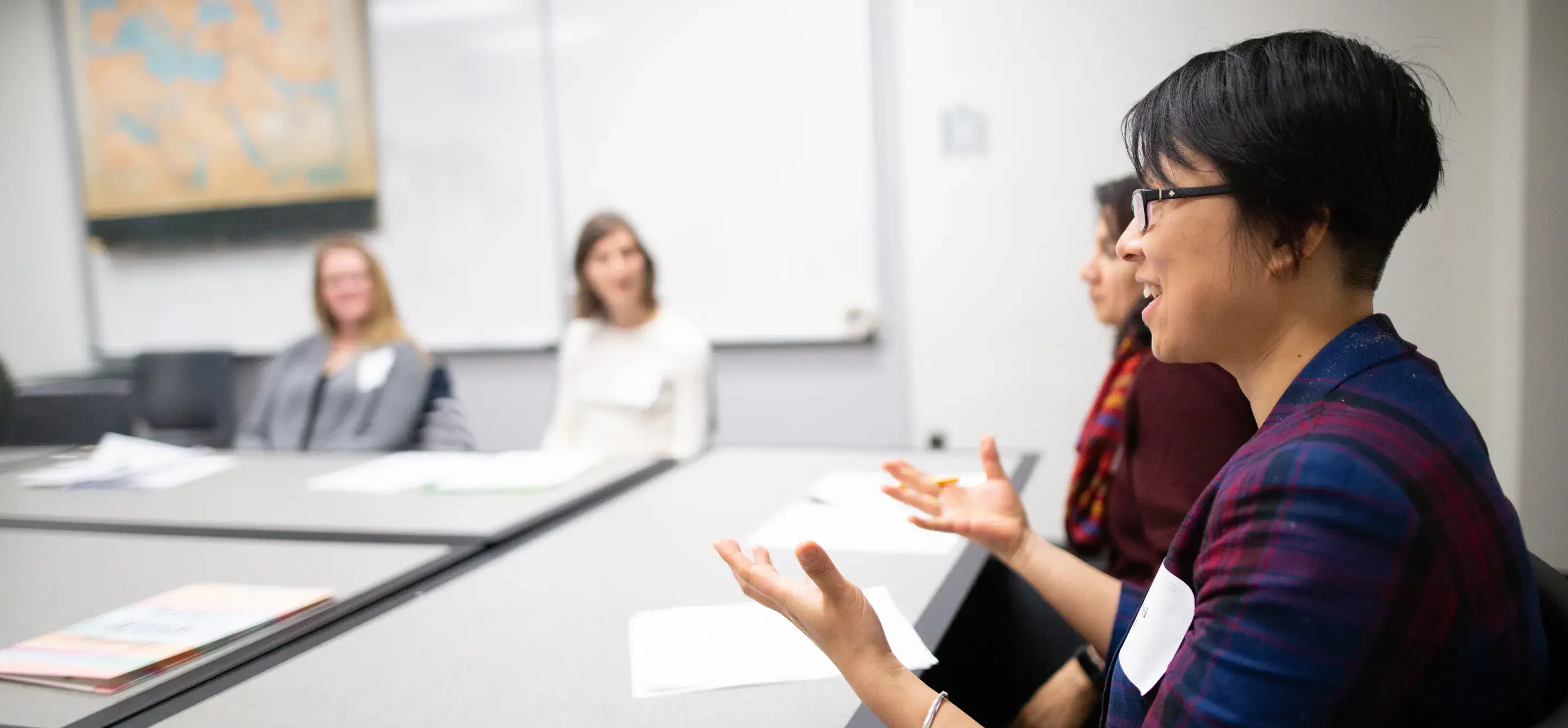 A woman with short hair and glasses speaks during a small group discussion in a classroom setting. She is animated, gesturing with her hands while speaking. Three other participants, two women and one person partially visible, are seated across the table, listening attentively. The room has whiteboards and a map on the wall, suggesting an academic or professional environment.