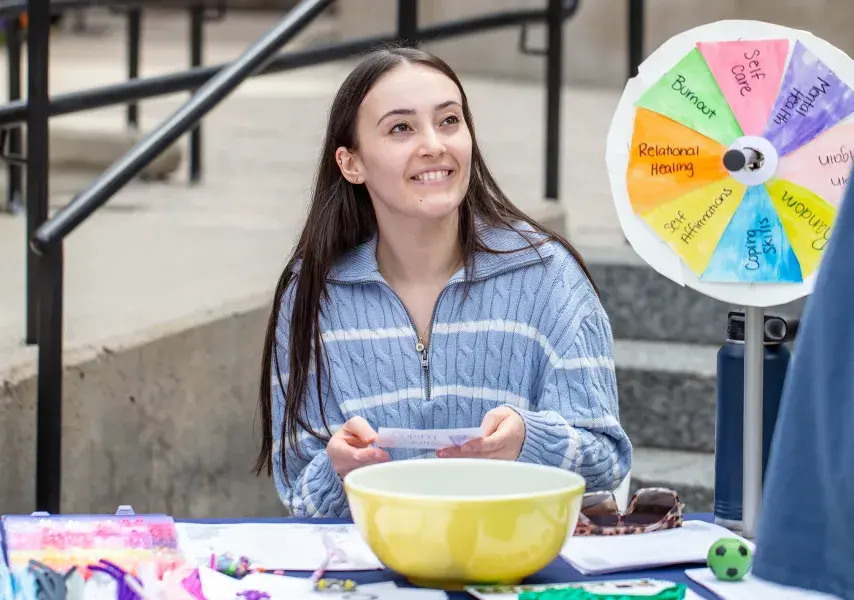 A friendly volunteer at a wellness fair engages with a visitor. She sits at an activity table featuring a colorful spinner wheel with positive themes and smiles as she holds a piece of paper, creating a welcoming and interactive atmosphere.