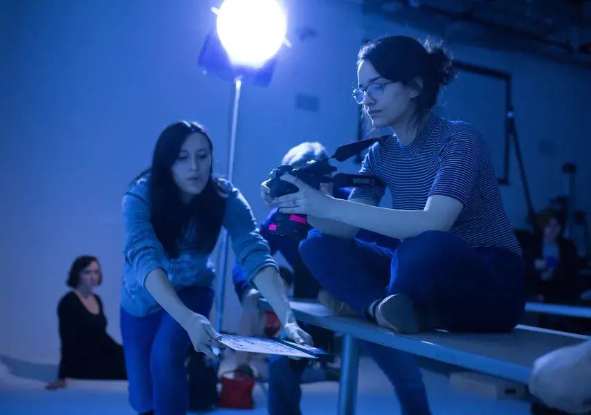 A behind-the-scenes view of a student photoshoot. Under the dramatic blue glow of a studio light, a photographer composes her shot while an assistant helps, demonstrating a creative and collaborative process.
