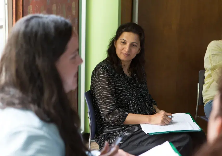 Two women are shown in a conversation during a meeting. The focus is on one woman who is actively listening, while the back of the head of the woman speaking is visible in the foreground.