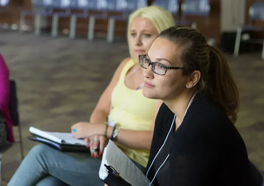 A close-up of a female attendee at a workshop, wearing glasses and a black shirt. Her rapt attention on an off-camera speaker conveys a sense of deep engagement and learning.