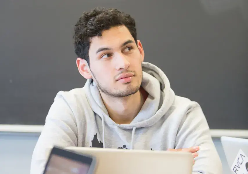 A young person in a light gray hoodie sits at a table in a classroom with a focused and contemplative expression, looking slightly upward. An Apple laptop is open in front of them, and two other laptops are visible in the foreground. The background features a chalkboard, indicating a school or college setting.